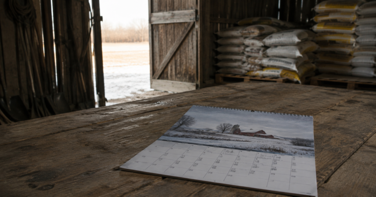 Winter farm calendar on a wooden table with feed sacks visible through an open barn door.